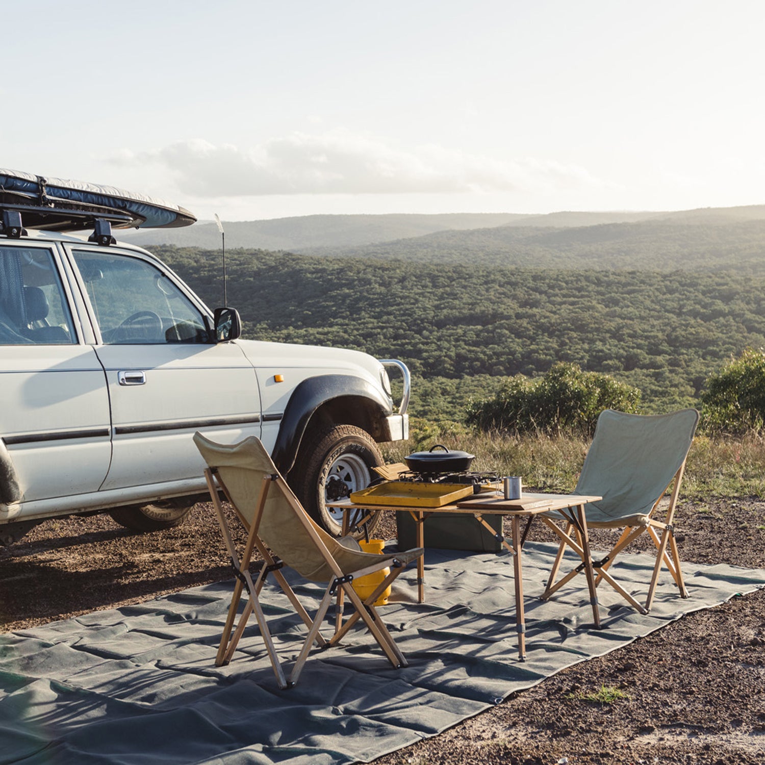 Camping setup with chairs, table, and a vehicle overlooking a scenic, green landscape.